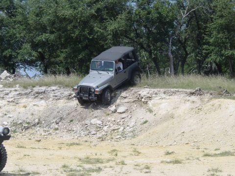 1973 Ford Bronco - A silver Jeep Wrangler navigates a challenging off-road terrain with trees in the background.