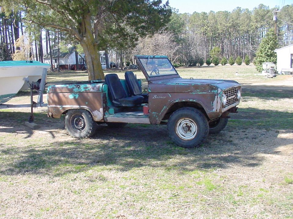 Rusty Ford Bronco with no top, sitting in a grassy yard next to a boat on a trailer