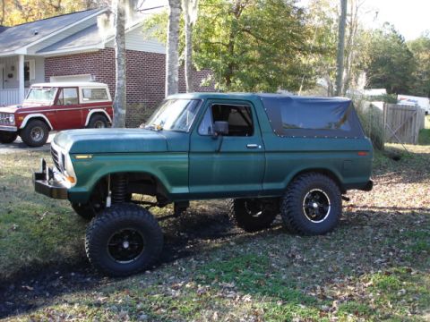 1979 Ford Bronco - Two Ford Broncos, one green and lifted, the other red, parked in a grassy area near a house.