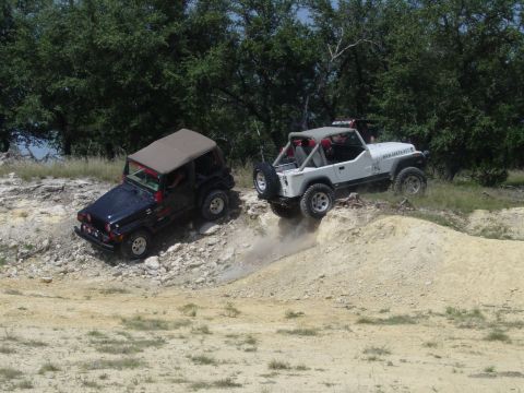 1973 Ford Bronco - Two Jeeps navigate a challenging off-road course, kicking up dust as one becomes airborne.