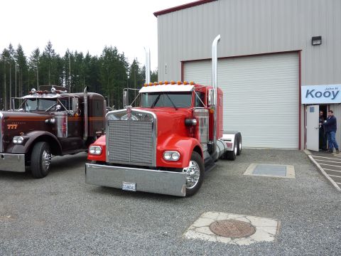 1981 Kenworth W900A - A bright red 1981 Kenworth W900A semi-truck is parked in front of a building, with another vintage truck to its left.