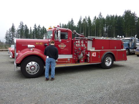 1981 Kenworth W900A - A red fire truck is parked on a gravel lot with trees in the background. Another truck is visible to the right.