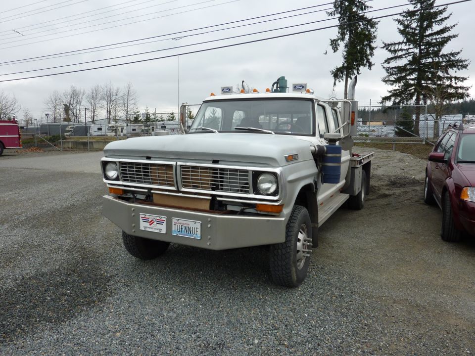 Front view of a 1970 Ford F-Series flatbed truck with a custom bumper and dual rear wheels. A red sedan is parked to the right