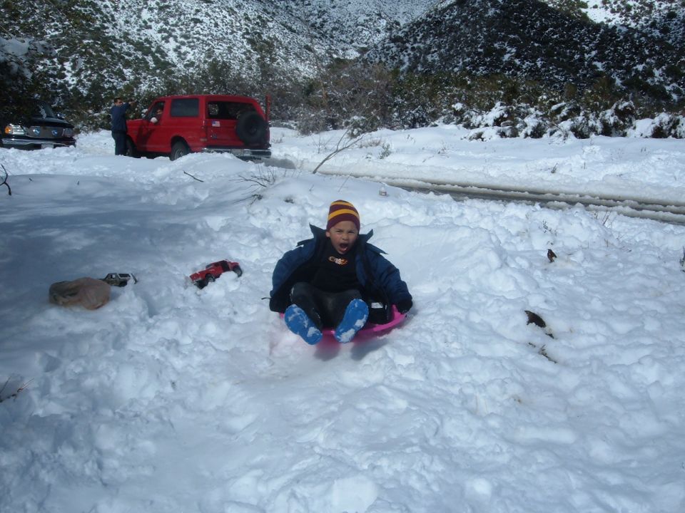 Child sledding on snow with a red Ford Bronco parked on the road behind