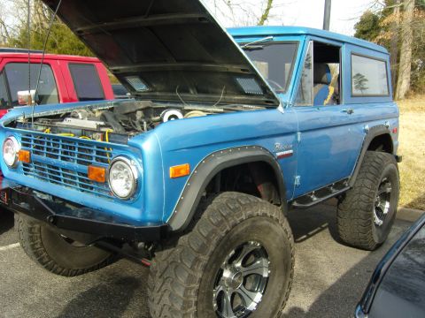 2007 Ford Mustang - A blue Ford Bronco is parked with its hood up, revealing the engine bay and a red truck behind it.