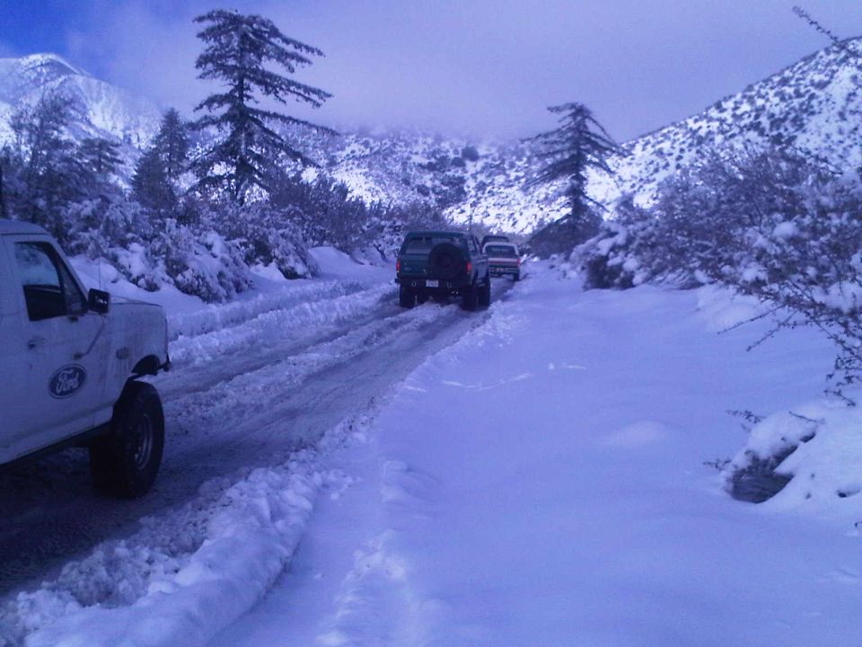 White Ford truck in foreground, two SUVs behind on snowy road, mountains and trees in background