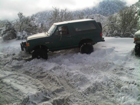 1986 Ford Bronco - A green 1986 Ford Bronco struggles in deep snow, with another vehicle nearby in the snowy terrain.