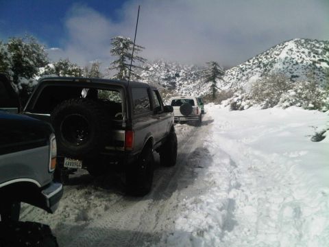 1986 Ford Bronco - Several vehicles navigate a snow-covered mountain road, with a dark-colored Ford Bronco in the foreground.