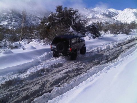 1986 Ford Bronco - A black 1987 Ford Bronco navigates a snow-covered mountain road, kicking up slush as it drives.