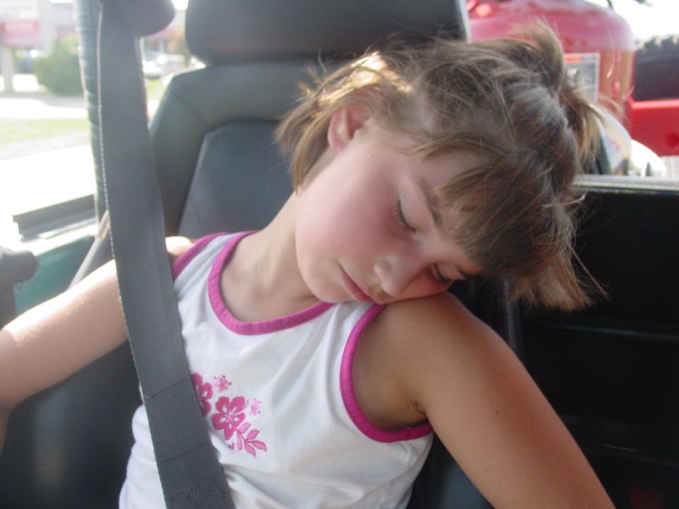 Young girl sleeping with her head on her shoulder, wearing a white tank top with pink trim and floral pattern. Seatbelt is visible