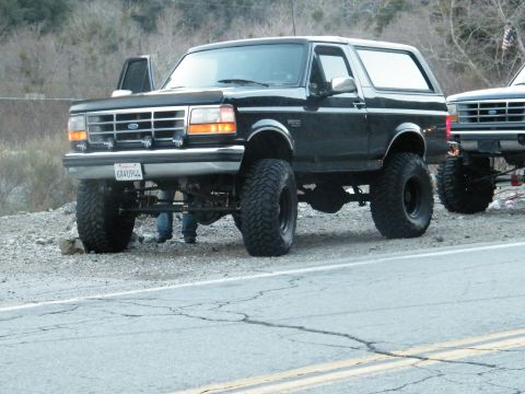 1982 Ford Bronco - Modified black Ford Bronco with large tires sits on a gravel shoulder next to a road.