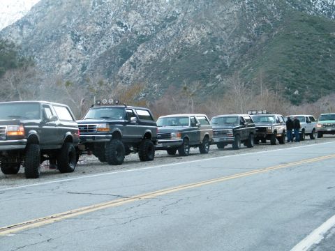 1982 Ford Bronco - A line of modified Ford Broncos parked on the side of a road, with a mountainous backdrop.