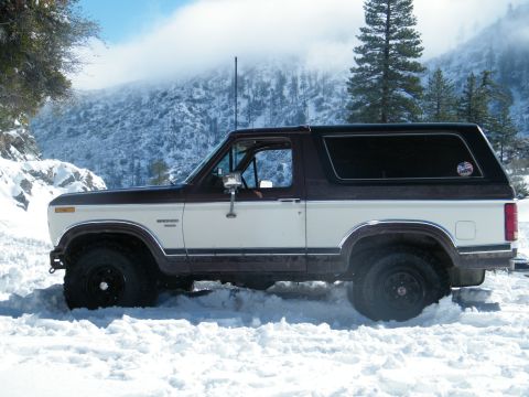 1982 Ford Bronco - A two-tone brown and white Ford Bronco navigates through deep snow in a mountainous, wooded area.