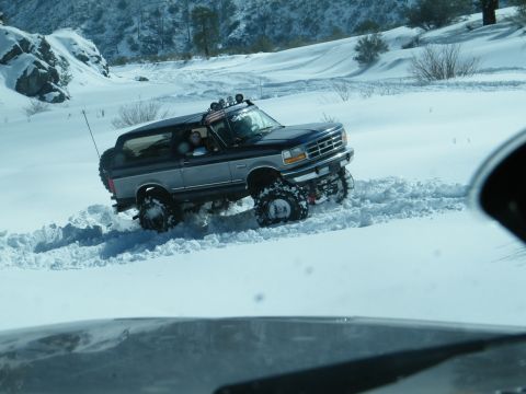 1982 Ford Bronco - This SUV navigates a snowy mountain pass, its off-road tires digging into the deep snow. The vehicle appears to be a 1992-1996 Ford Bronco.