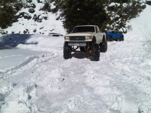 1986 Ford Bronco - A lifted 1987 Ford F-150 pickup truck navigates a snowy terrain, with another vehicle visible in the background.