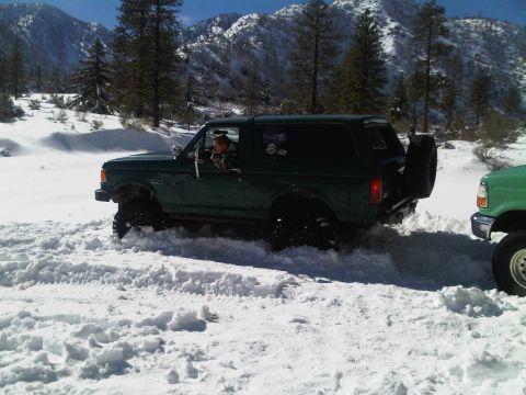 1986 Ford Bronco - A green Ford Bronco navigates a snowy mountain road, with another vehicle visible to the right.