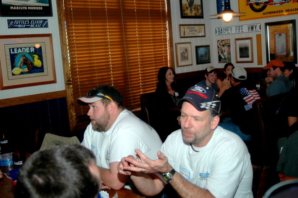 Men wearing t-shirts and caps sit at tables in a restaurant with wooden blinds and framed pictures on the wall