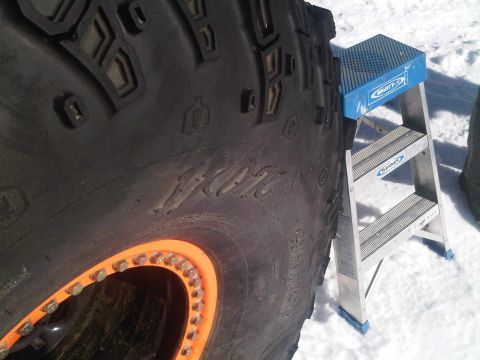 1986 Ford Bronco - Close-up of a large off-road tire with an orange beadlock wheel, next to a step stool in a snowy environment.