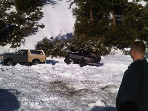 1986 Ford Bronco - Two Ford Broncos appear stuck in a snowy, wooded area, with one vehicle showing off-road modifications.