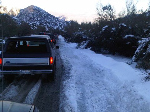 1986 Ford Bronco - Several SUVs are driving in a convoy on a snow-covered road with mountains in the background.