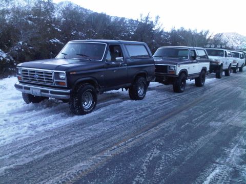 1986 Ford Bronco - A convoy of Ford Broncos drives down a snow-covered road, navigating the winter conditions.