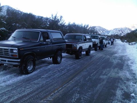 1986 Ford Bronco - A line of Ford Broncos drive down a snow-covered road in a mountainous area.
