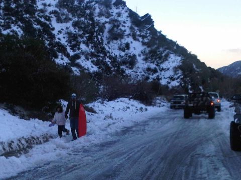 1986 Ford Bronco - A snowy mountain road scene with two people holding a sled, and several vehicles driving or parked.