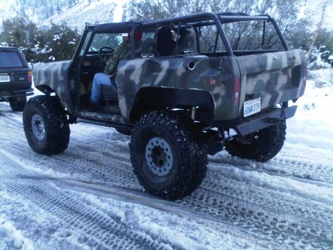 1986 Ford Bronco - An off-road vehicle with a camouflage paint scheme drives on a snowy surface, with another vehicle visible to the left.