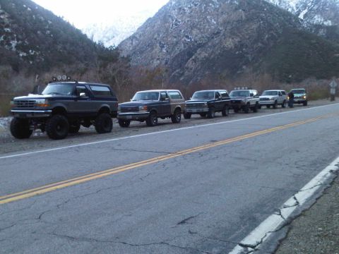 1986 Ford Bronco - Several Ford Broncos and other SUVs are lined up on the side of a road, with a mountainous landscape behind them.