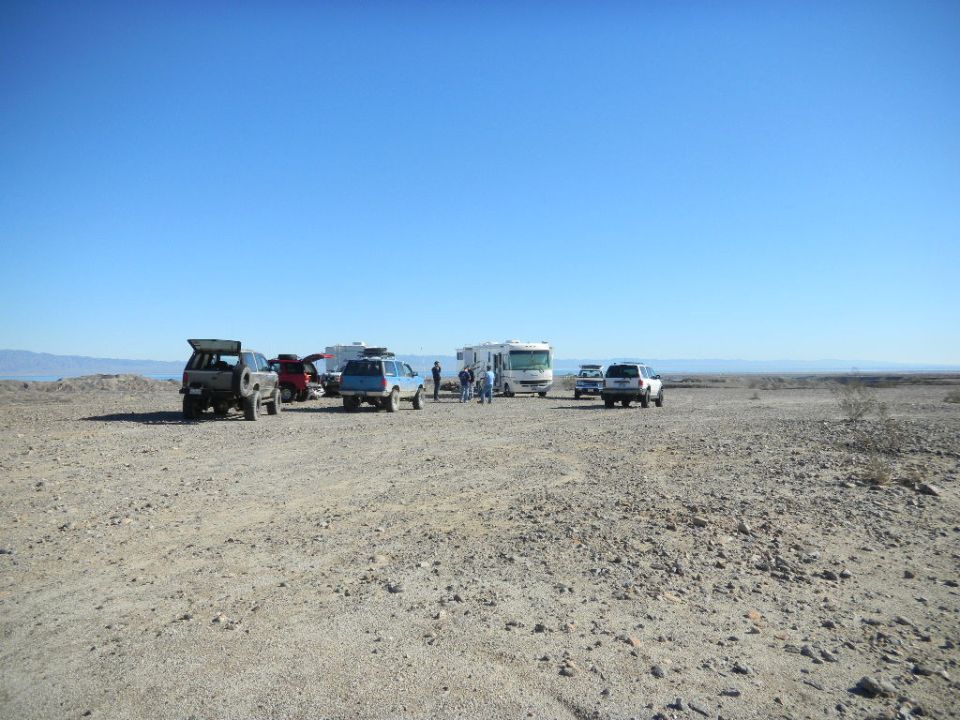Desert scene with multiple SUVs, trucks, and RVs parked on a dirt lot under a clear blue sky