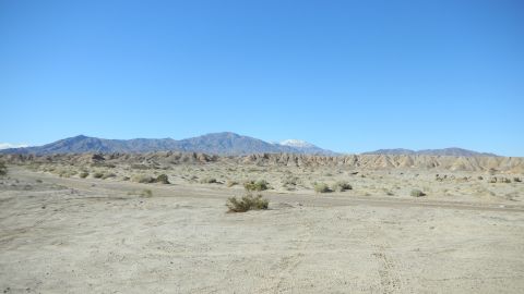 1997 Ford Ranger - Wide open desert landscape with a sandy wash marked by tire tracks and scattered creosote brush, backed by eroded badlands ridges and a sno…