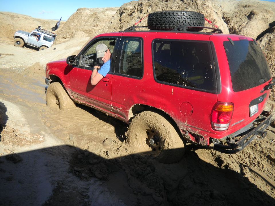 Red Ford Explorer stuck in mud, driver looking out window. Silver SUV in background. Spare tire on roof rack