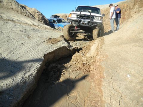 1997 Ford Ranger - Two trucks navigate a challenging off-road course with steep dirt inclines and a muddy ditch.