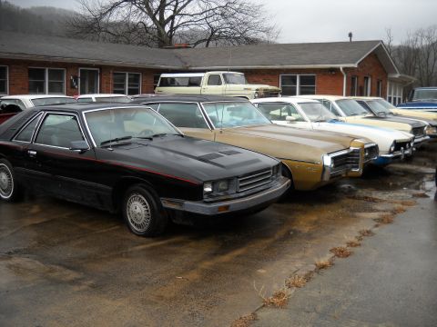 1982 Ford Bronco - Several older model cars are lined up in front of a brick building on a cloudy day.