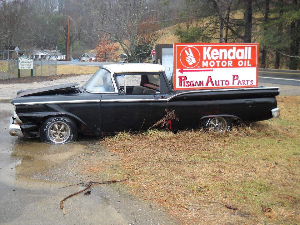 Black 1959 Ford Ranchero with a white roof, hood up, parked on grass next to a Kendall Motor Oil sign