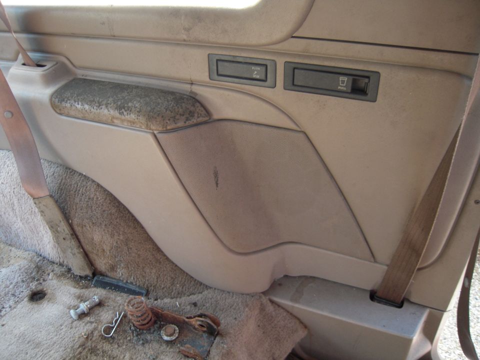 Worn tan interior door panel with armrest and window switches, parts on floor below