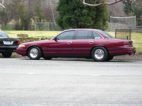 1992 Ford Crown Victoria - A maroon sedan is parked on the street next to another vehicle, with trees and a fence in the background.