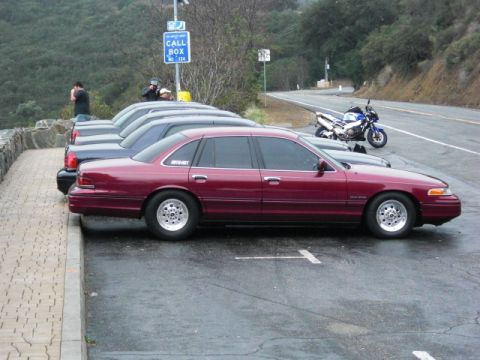 1992 Ford Crown Victoria - A line of sedans, including a maroon 1992 Ford Crown Victoria, are parked near a call box on a cloudy day.
