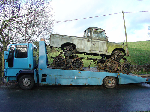 Silver track-converted 4x4 loaded on a blue flatbed recovery truck, side view outdoors.