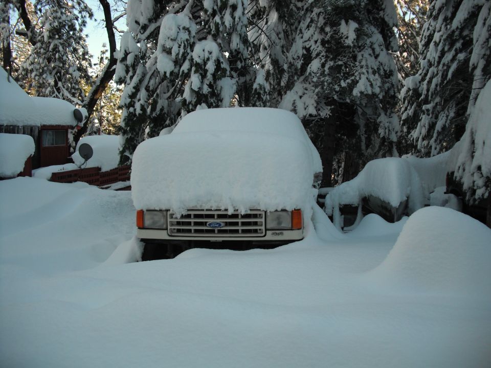 Front view of a 1991 Ford Bronco buried in snow, parked outside a house with snow-covered trees