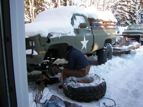 1990 Chevrolet Suburban - A person works on the front wheel of a snow-covered olive green pickup truck with a white star emblem.