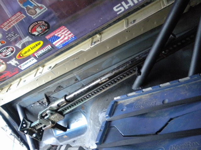 Hi-Lift jack mounted in a bare truck bed with roll cage tubing and decal-covered rear glass