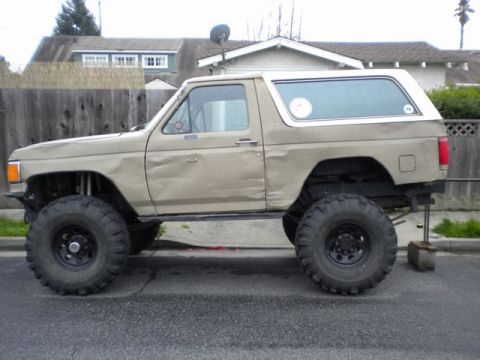 1989 Ford Bronco - This lifted Ford Bronco has large off-road tires and visible damage to its doors.