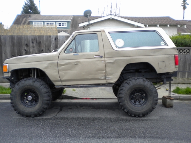 Tan lifted Ford Bronco on street with white rear top removed and propped on a stand