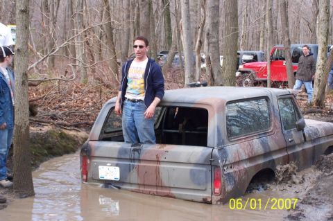 1979 Ford Bronco - This camouflaged SUV is deeply stuck in the mud, with several people observing the scene in a wooded area.