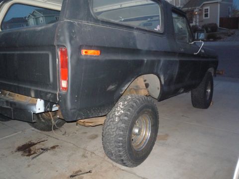 1979 Ford Bronco - Three-quarter rear view of a flat-black lifted Ford Bronco sitting in a driveway with the rear wheel removed and the axle on wood blocks, e…