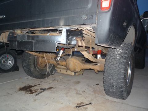 1979 Ford Bronco - Low rear-quarter shot under a flat-black lifted Ford Bronco showing the exposed dirt-caked solid rear axle housing, leaf springs, red-bodie…