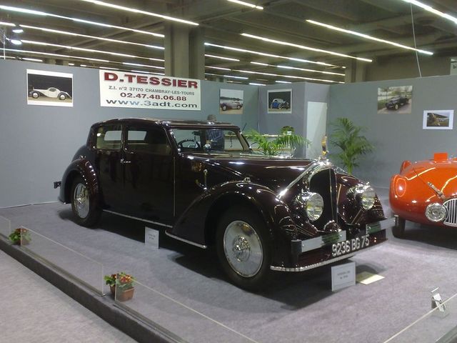 Maroon prewar luxury sedan on a show stand beside an orange roadster