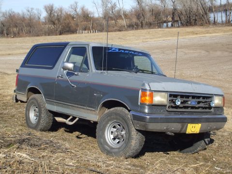 1989 Ford Bronco - A two-tone Ford Bronco is shown from a three-quarter front angle, parked outdoors on a dry, grassy field.