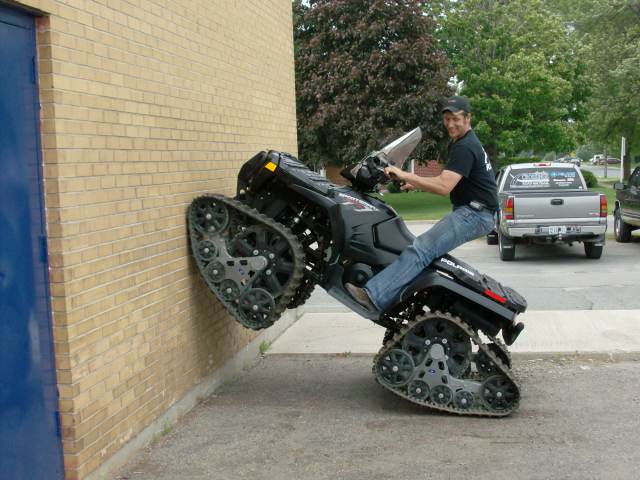 Black tracked ATV doing a wheelie against a brick wall with a rider on top